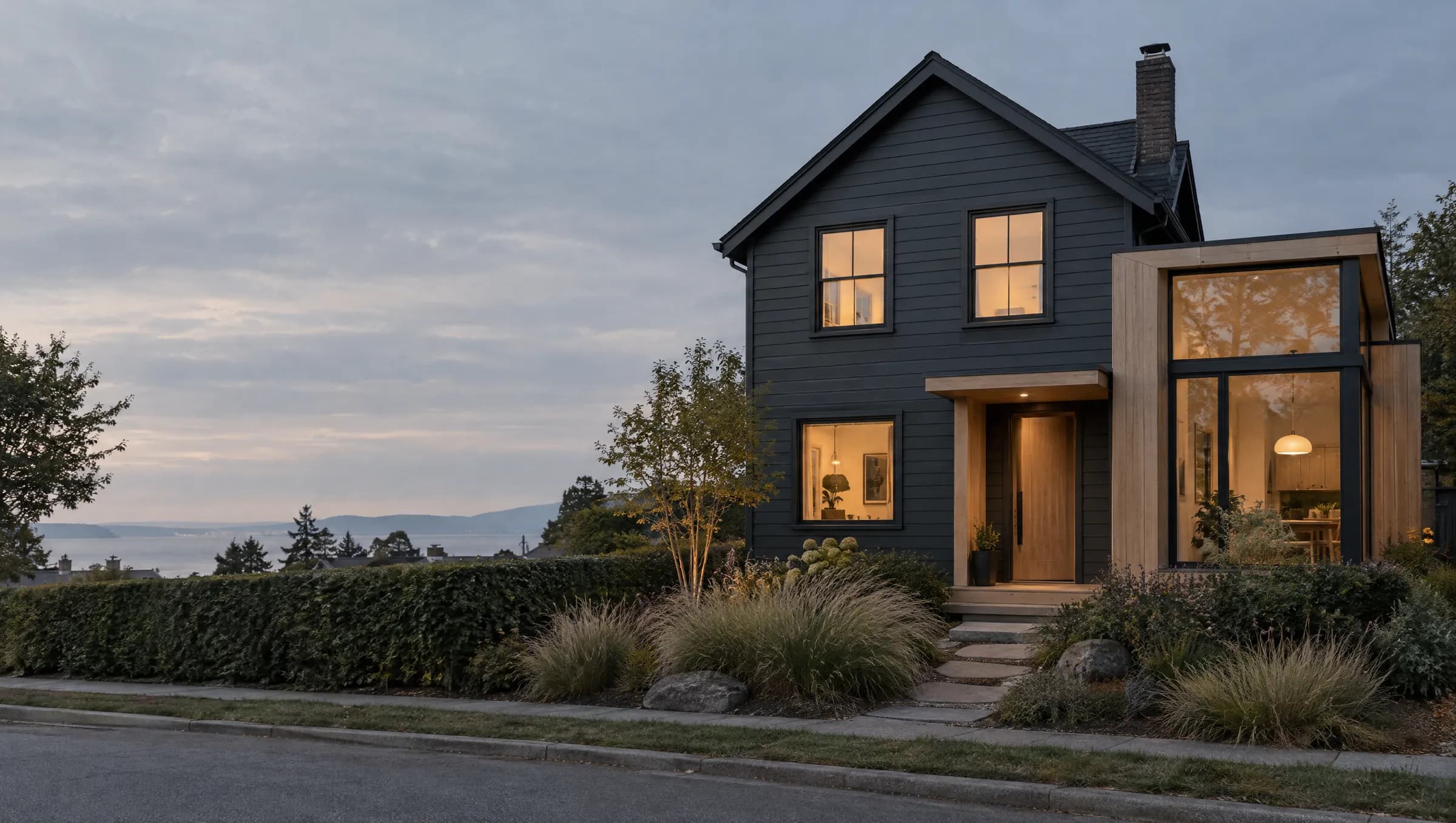 A renovated charcoal clapboard house with a white-oak extension at dusk, its windows lit, the sea behind.